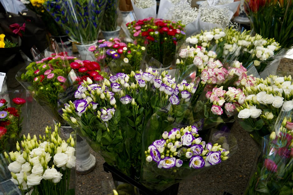 A vibrant display of fresh flower bouquets arranged on a table, including white and purple Lisianthus, pink and white Eustoma, and red and pink everlasting daisies, all wrapped in clear cellophane. Th