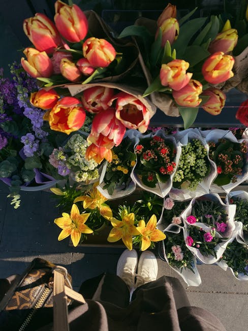 A variety of fresh flowers arranged in bouquets on display at a florist shop. The bouquets feature vibrant tulips with yellow and pink petals, complemented by clusters of purple, white, and red flower