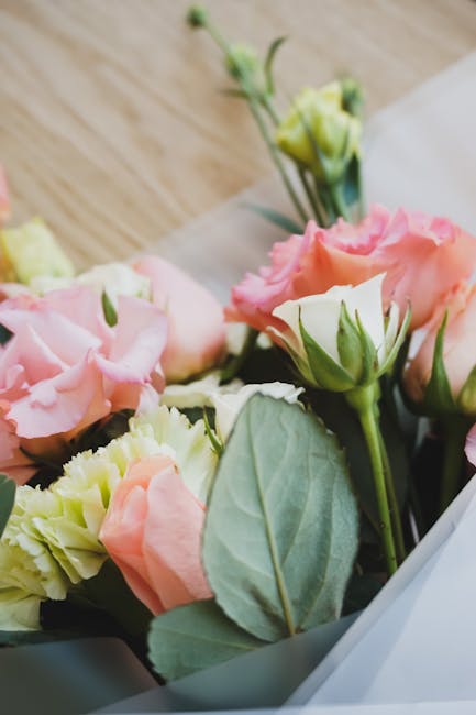 A close-up view of a bouquet featuring light pink and soft peach lisianthus flowers with ruffled petals, accented by fresh green leaves and stems. The flowers are arranged in a casual, natural style o