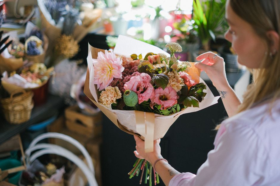 A woman with long blonde hair, wearing a light pink blouse, holds a large bouquet of fresh flowers in a florist shop. The bouquet features soft pink roses, cream-colored carnations, pale pink eustoma,