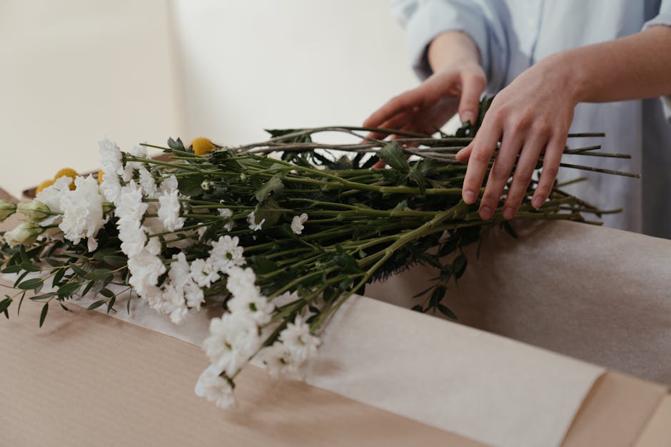A person’s hands are carefully arranging a bouquet of fresh white and yellow flowers, including carnations and daisies, on a light-colored wooden surface. The bouquet features vibrant yellow blooms 